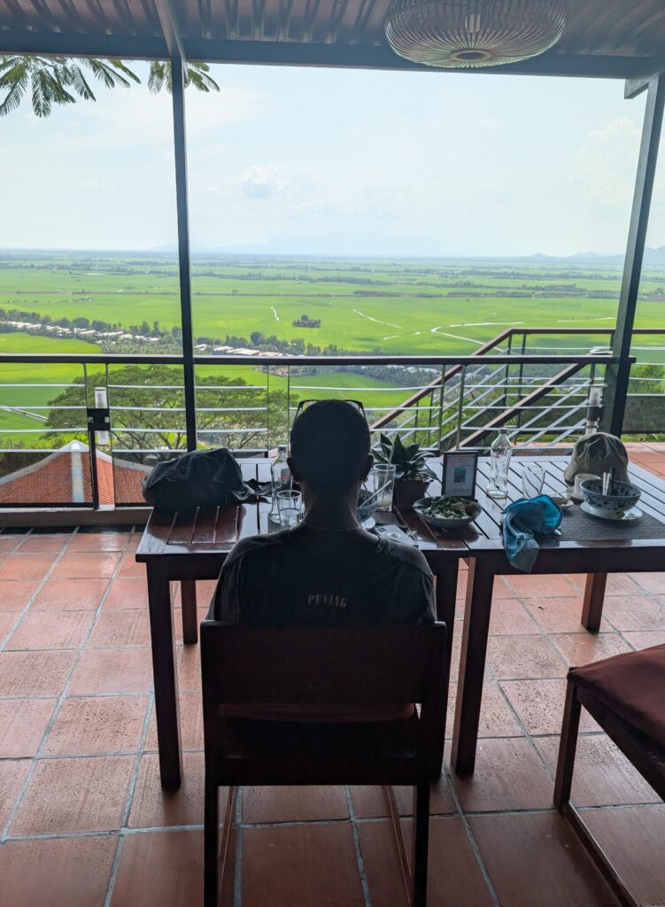 Mary-Ann Sat at a table eating Pho and looking across an expanse of Vietnamese countryside.