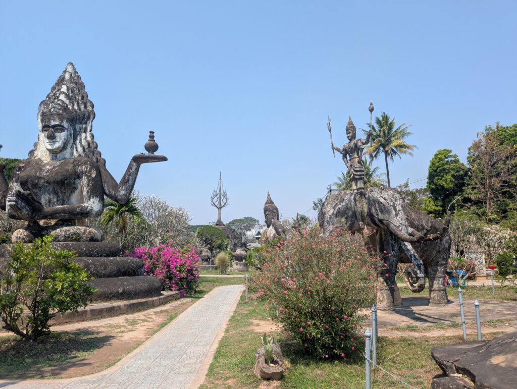A view of the Buddha park, showing multiple buddhist statues, pink flowers and blue sky.