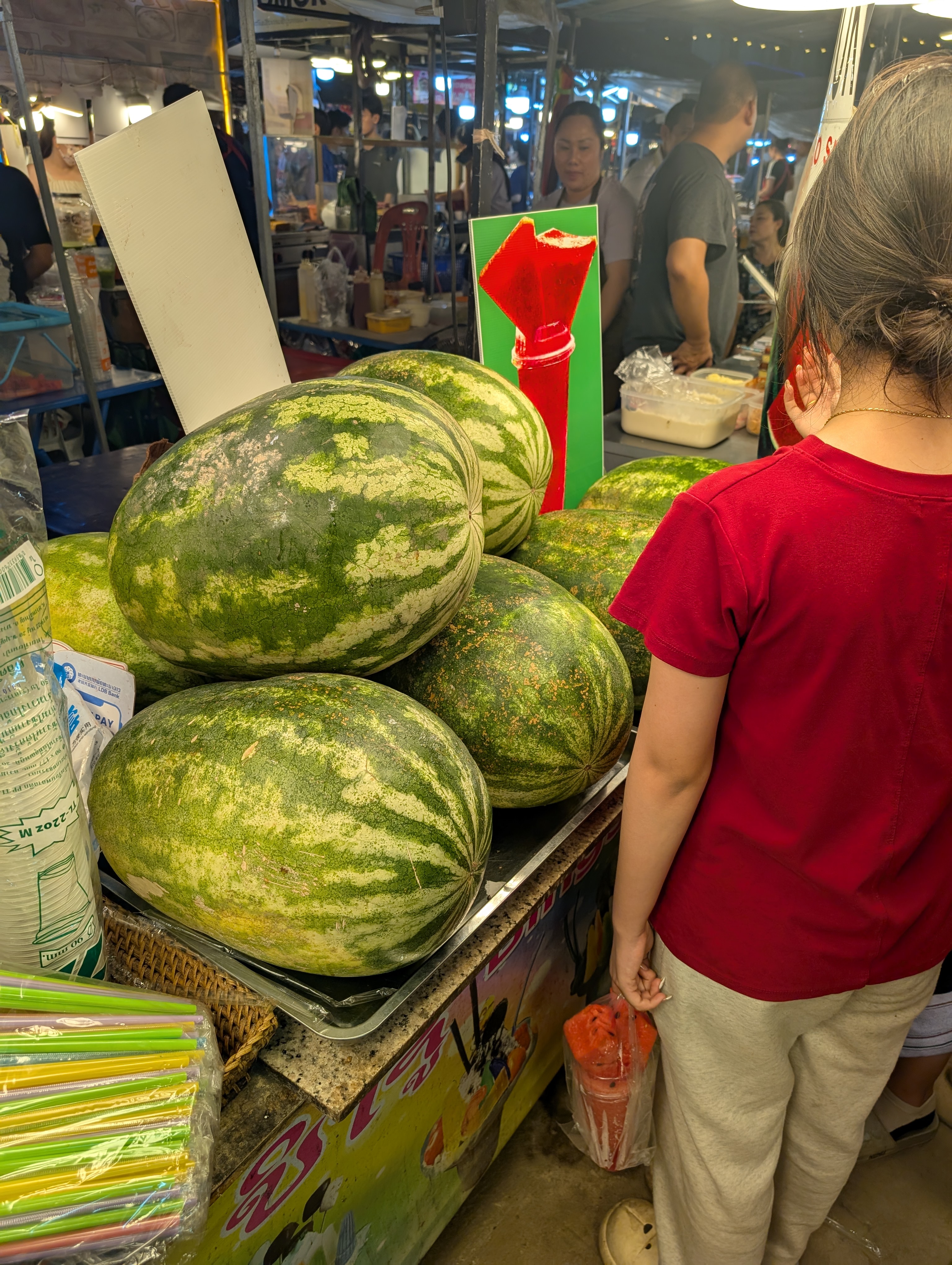 A picture of a pile of huge watermelon. They are the same size as the top half of the lady standing next to them.