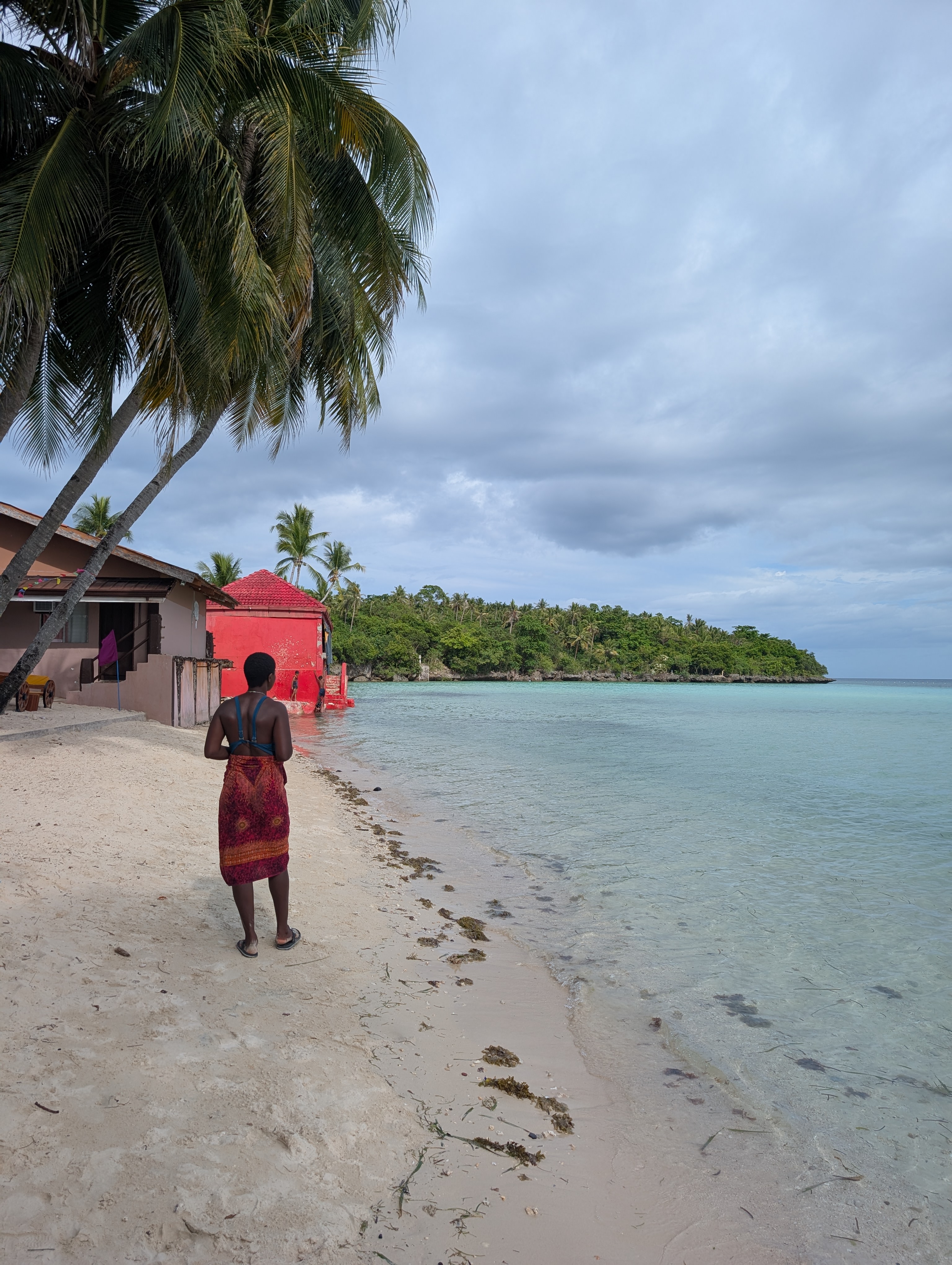 Mary-Ann standing on a beach, with some local houses and a green headland in the distance.