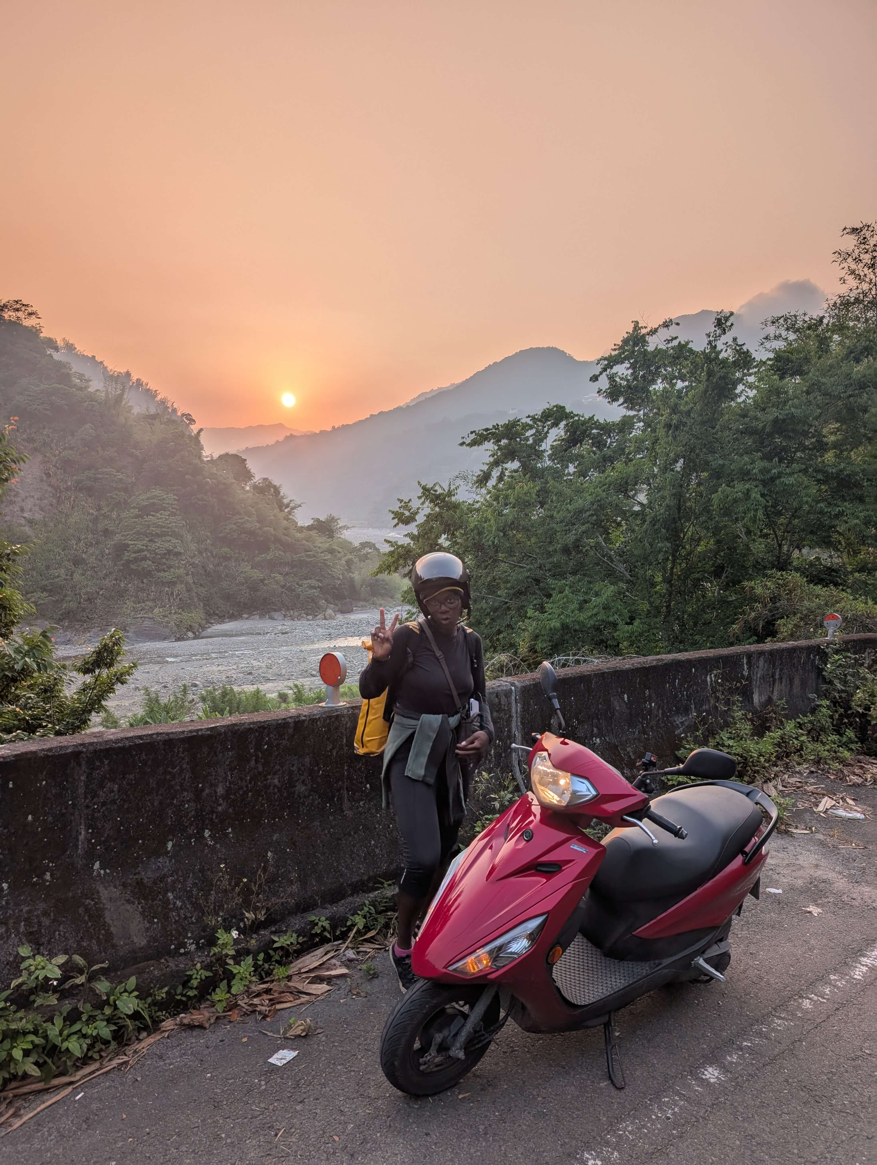 Mary-Ann stood next to the moped infront of a sunset