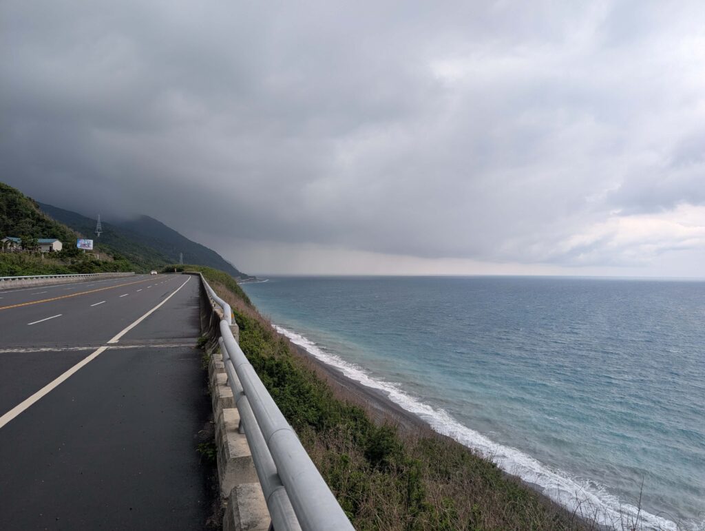 A shot looking up the east coast coastal rooad. The sea is blue, the road is smooth and there are thick rain clouds rolling over the mountain.