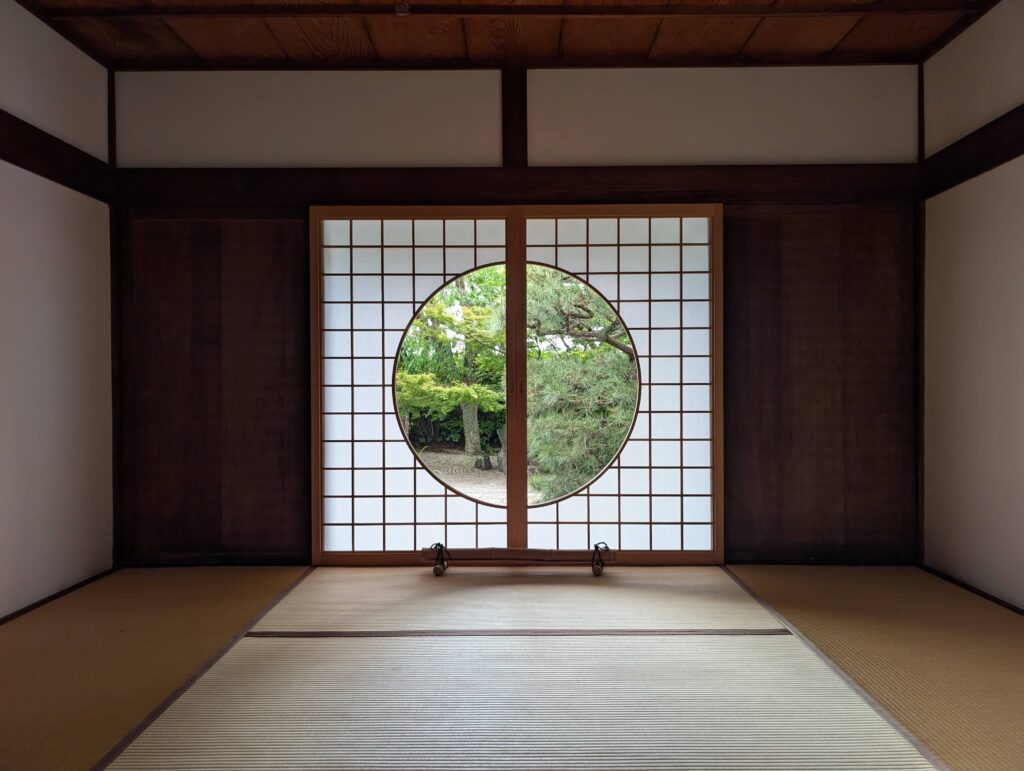 An image of a Japanese room with rattan wood floor and round window looking out into a garden.